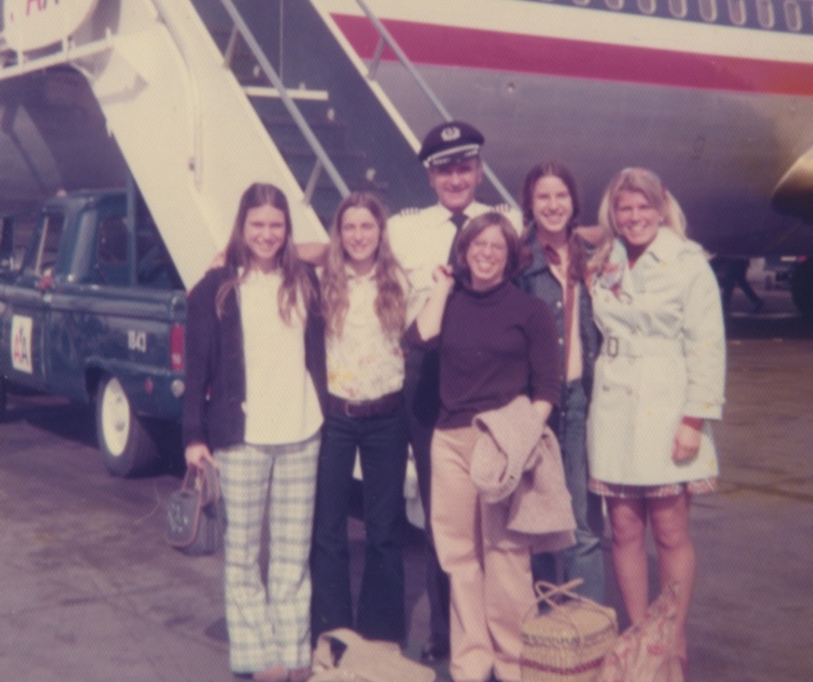 Swim coach Sue Davis (far right) takes her team on a training trip. They stand with a pilot in front of a set of stairs attached to an airplane. 